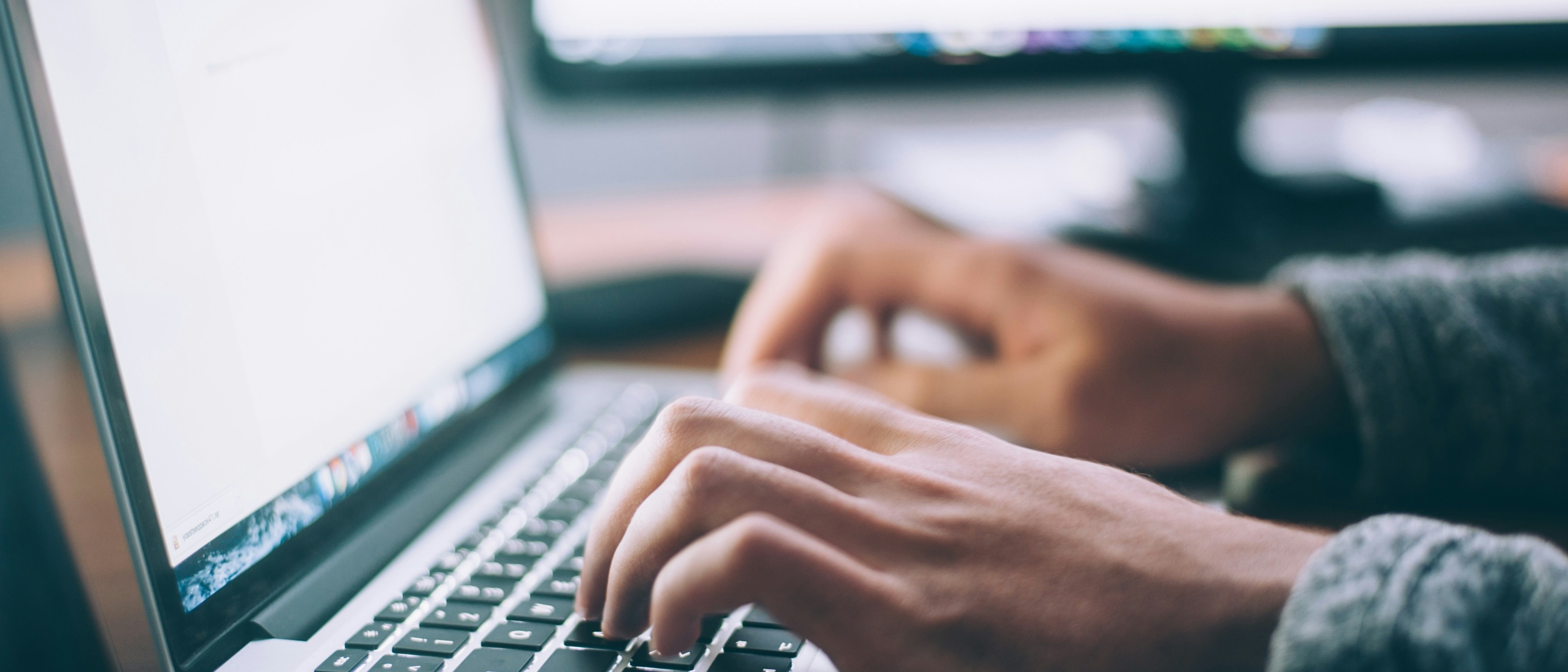 Hands typing on a laptop keyboard, with a blurred computer monitor in the background